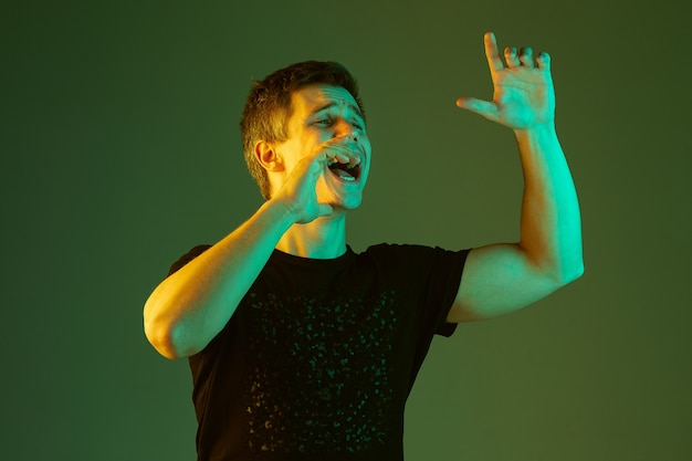 Calling somebody, screaming. Caucasian man's portrait isolated on green studio background in neon light. Beautiful male model in black shirt. Concept of human emotions, facial expression, sales, ad.