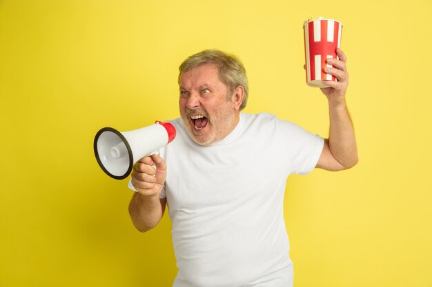 Calling with mouthpeace, holds popcorn. Caucasian man portrait on yellow studio background. Beautiful male model in white shirt. Concept of human emotions, facial expression, sales, ad. Copyspace.