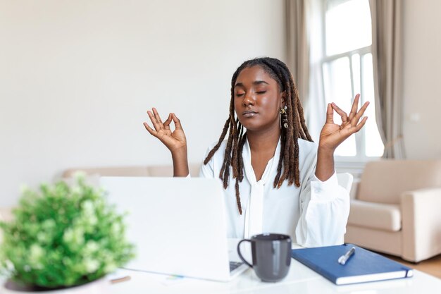 Calm African female executive meditating taking break at work for mental balance mindful businesswoman feeling relief and no stress doing yoga at work ignoring avoiding stressful job