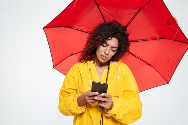 Calm african woman in raincoat hiding under umbrella and writing message on her smartphone over white background