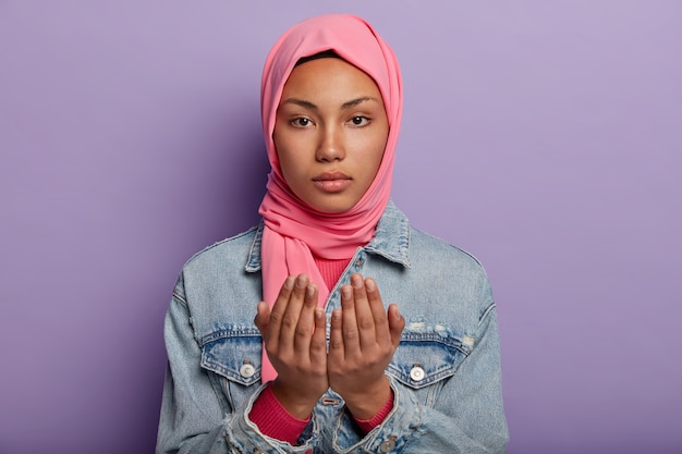 차분한 매력적인 아랍 여성이기도하는 제스처에 손바닥을 유지하고 분홍색 히잡과 데님 재킷을 입습니다.