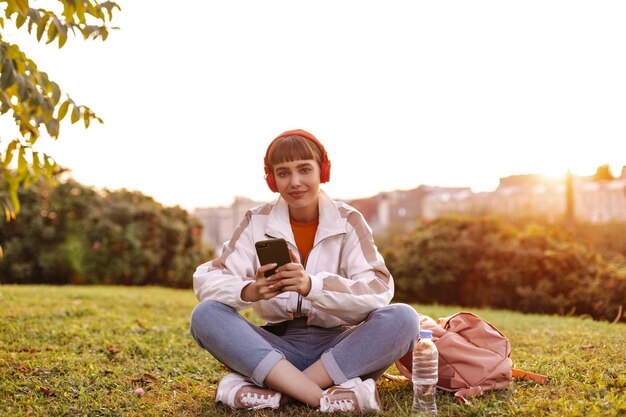 Спокойная брюнетка в джинсовой белой куртке сидит на траве во время заката, держит смартфон и слушает музыку в наушниках