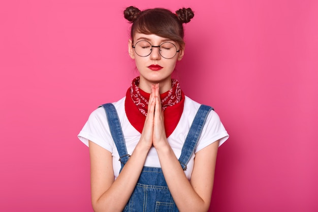 calm Caucasian female keeps eyes closed, prays while standing against pink wall, asks God for something important, wears white t shirt, denim overalls, bandana on neck and glasses.