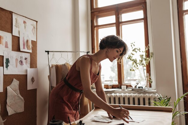 Calm charming shorthaired woman in linen red dress with black belt work as fashion designer in cozy office and paint garment pattern