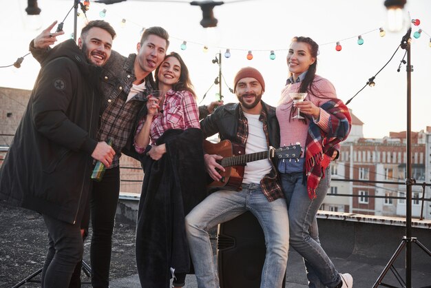 Calm and cheerful. Party at the rooftop. Five good looking friends that posing for the picture with alcohol and guitar