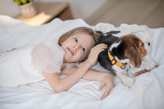 Calm cute long-haired Caucasian female child lying on the bed next to her four-legged friend