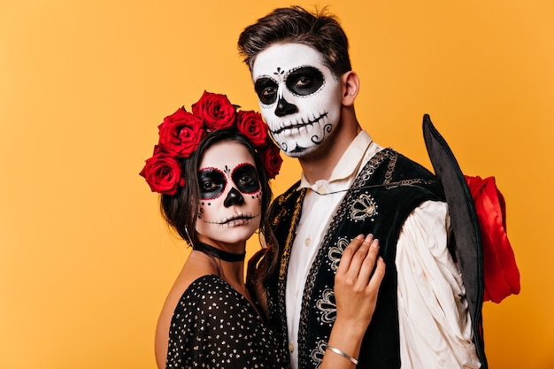 Calm guy and dark-haired girl. Woman with crown of roses embraces Mexican man with painted face.