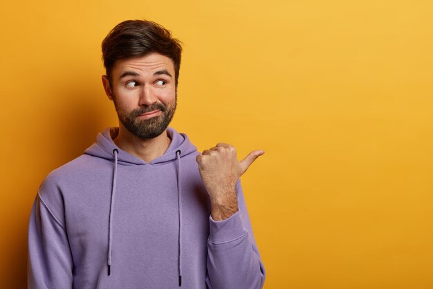 Calm handsome bearded Caucasian man with curious expression points thumb aside on blank space, demonstrates good promo, place for your advertising, wears hoodie, poses over yellow  wall