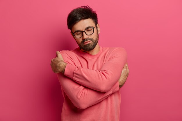 Calm handsome bearded man cuddles himself, feels comfort and coziness, recalls romantic date with girlfriend, closes eyes and tilts head, wears sweater and spectacles, isolated on pink wall