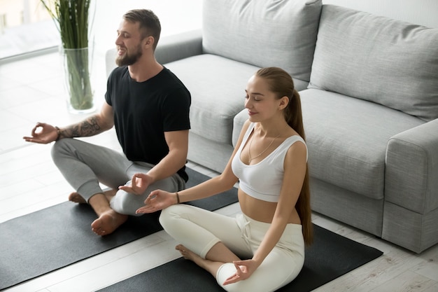 Calm millennial couple meditating in lotus position at home