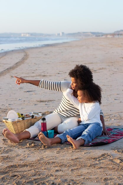 Madre e figlia calma che fanno picnic sulla spiaggia madre e figlia afroamericane in abiti casual seduti su una coperta, discutendo del tramonto. famiglia, relax, concetto di natura