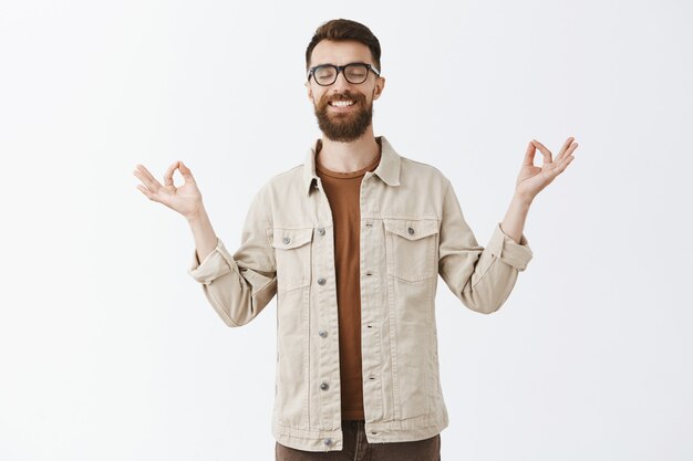 Calm patient bearded man in glasses posing against the white wall