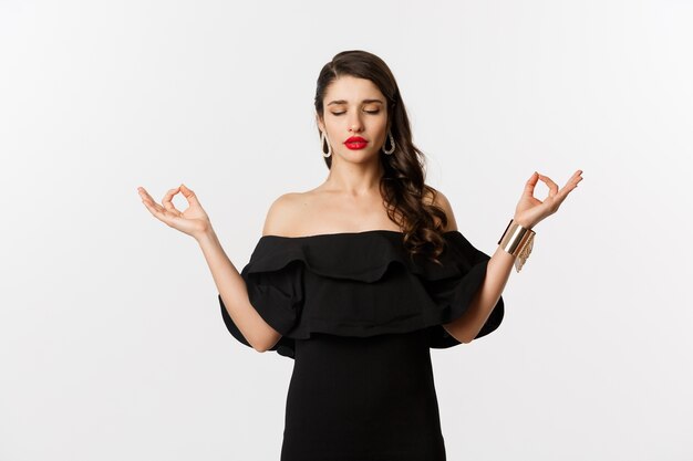 Calm and patient young brunette female meditating, standing in black dress with hands in zen gesture, standing against white background.