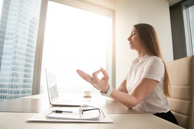 Calm peaceful businesswoman practicing yoga at work, meditating in office