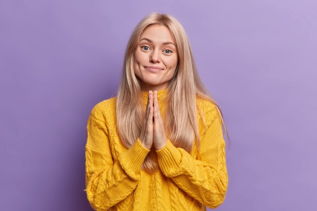 Calm pleased blonde young European woman presses palms together and asks for favor needs your help wears casual yellow sweater 