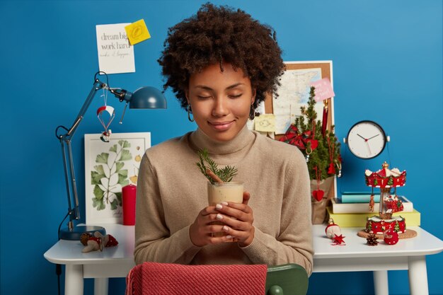 Calm relaxed African American woman poses with eggnog cocktail, sits with closed eyes at chair, wears brown turtleneck