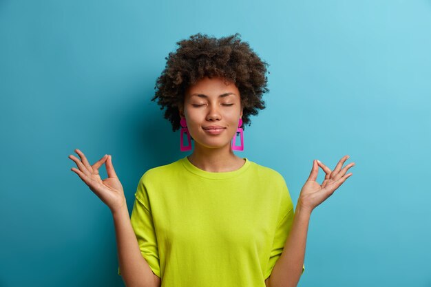 Calm relaxed dark skinned woman stands in lotus pose, feels relieved, tries to concentrate in peaceful atmosphere, wears casual green clothes, isolated on blue wall. Body language concept