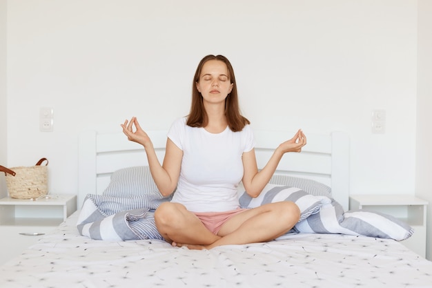 白いカジュアルなスタイルのTシャツとショートパンツを着て、蓮華座で明るい寝室のベッドに座って、ヨガの練習と瞑想をしている黒髪の穏やかなリラックスした女性。