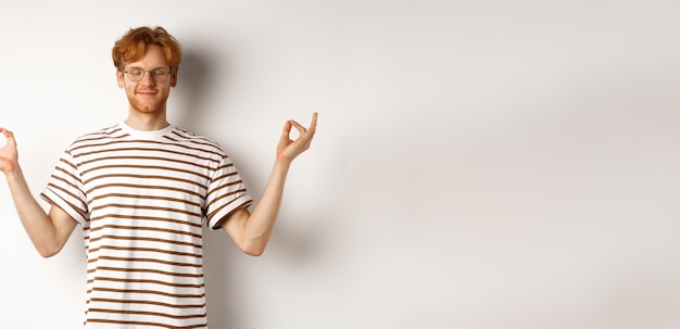 Free photo calm and relaxed young man with red messy hair spread hands sideways in mudra gesture and smiling pr