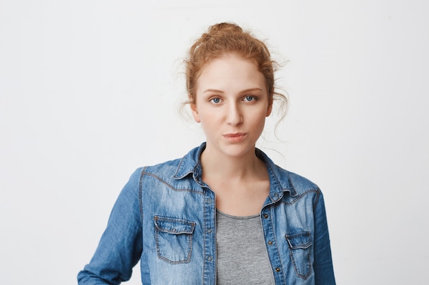 Calm serious caucasian ginger girl with hair combed in bun, expressing irritation or indifference while standing