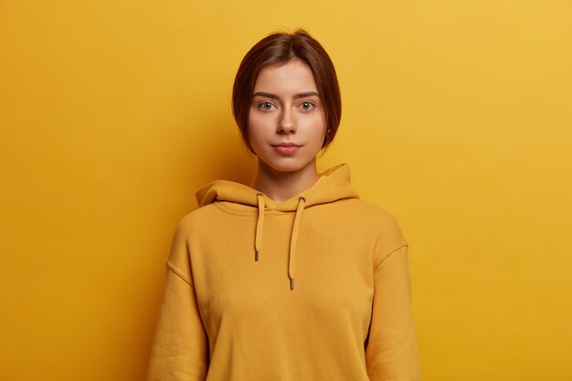 Calm serious green eyed European teenage girl looks straightly , has casual talk, discusses everyday events, has straight combed hair, wears hoodie, isolated over bright yellow wall
