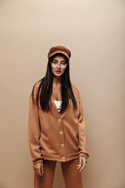 Free photo calm serious woman in light brown stylish cashmere suit and cap looks into camera brunette longhaired girl in eyeglasses poses on isolated beige background