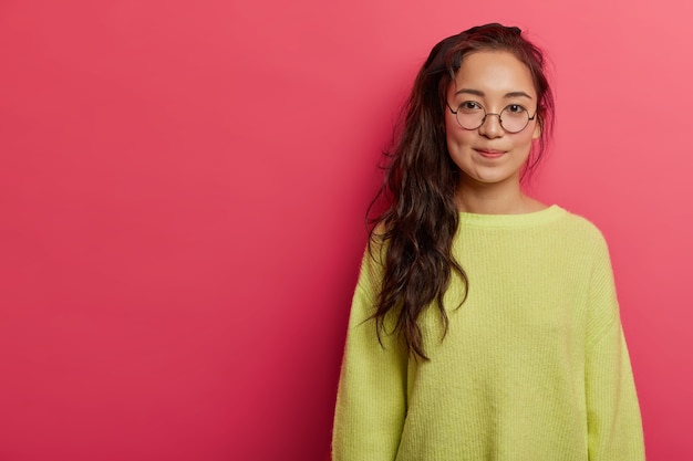 Calm serious woman with long pony tail, wears transparent glasses, green jumper, looks attentively at camera, poses in pink studio