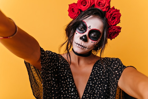 Calm slender girl makes selfie, showing off her image for Halloween. Indoor portrait of pretty model on orange wall