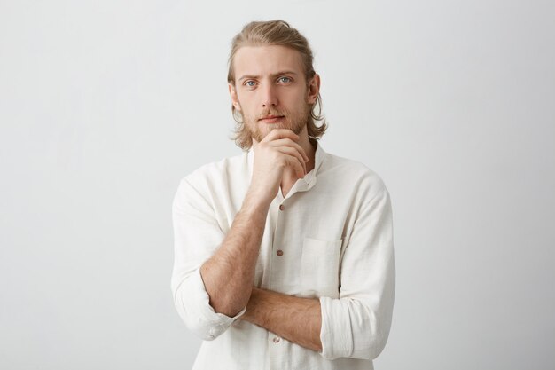 Calm successful european bearded male with fair hair, holding hand on chin