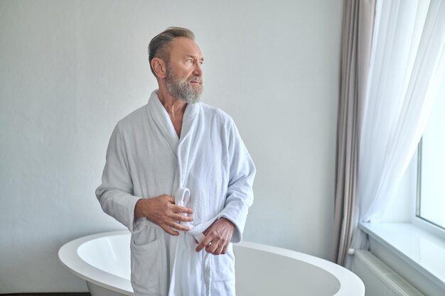 Calm thoughtful gray-haired bearded mustached mature man fastening the belt of his white terry bathrobe