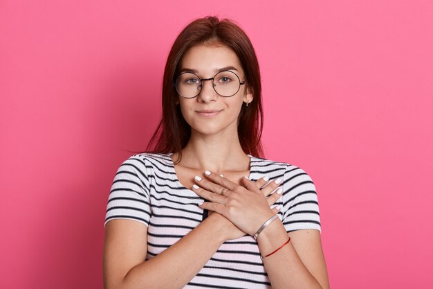 穏やかな女性は感謝し、愛を示し、彼女の胸に手を保ち、ストライプのTシャツとメガネを着用し、ピンクの壁に孤立したポーズをとります。