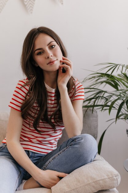 Calm woman in jeans and T-shirt is resting in apartment and sitting on soft sofa