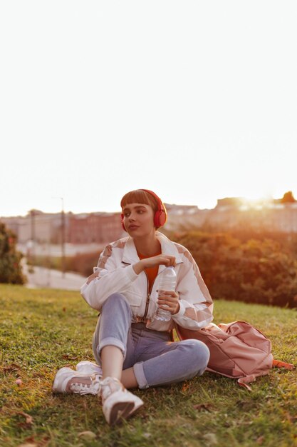 Calm woman sits on grass outside and holds water bottle Shorthaired girl in stylish jacket and jeans listens to music in headphones