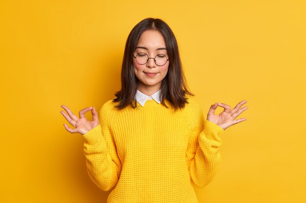 calm young Asian woman shows zen or okay sign meditates in good mood stands with eyes closed wears casual sweater and spectacles.