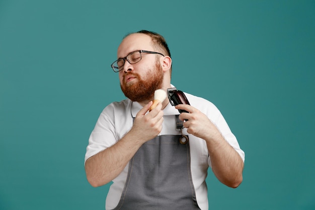 Calm young barber wearing uniform and glasses turning head to side holding shaving brush and hair trimmer trimming face with closed eyes isolated on blue background