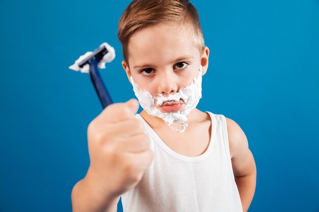 Calm young boy in shaving foam like man showing razor