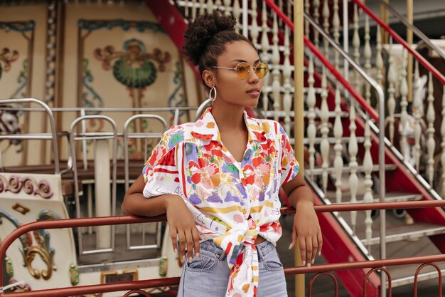 Calm young brunette curly woman in denim pants, colorful blouse and orange sunglasses looks away and leans on carousel outside