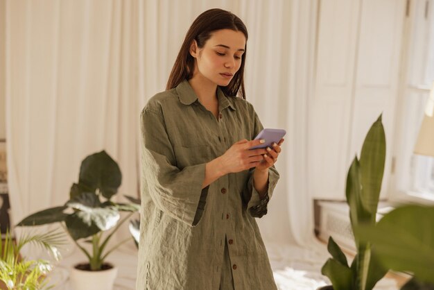 차분한 백인 여성이 실내에서 휴대전화 화면을 보고 긴 녹색 셔츠를 입은 갈색 머리는 소셜 네트워크 기술 장치 개념에서 SMS를 읽습니다.