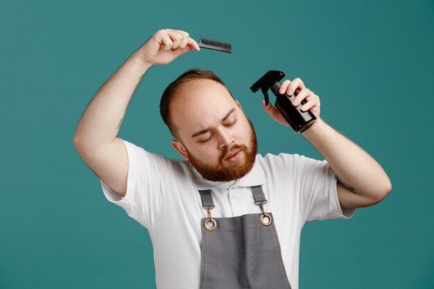 calm young male barber wearing white shirt and barber apron holding hair spray near head and teaser comb above head with closed eyes isolated on blue background