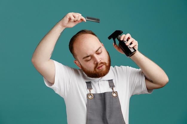 Calm young male barber wearing white shirt and barber apron holding hair spray near head and teaser comb above head with closed eyes isolated on blue background