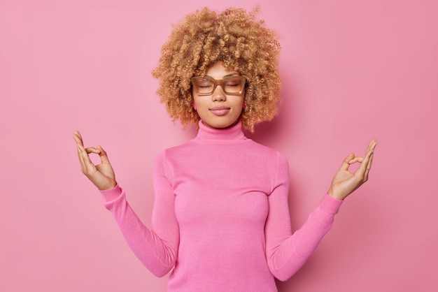 Calm young peaceful sound minded woman meditates with eyes closed makes om gesture tries to concentrate wears eyeglasses and turtleneck isolated over pink background makes yoga breathing practice