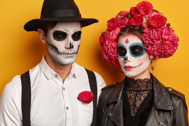 Calm young woman and man wears skull makeup, female in beautiful floral wreath, dressed in halloween holiday costumes, keep eyes shut, isolated over yellow studio background.