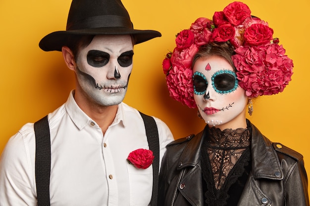 Спокойная молодая женщина и мужчина в макияже черепа, женщина в красивом цветочном венке, одетая в праздничные костюмы на Хэллоуин, держат глаза закрытыми, изолированные на желтом фоне студии.