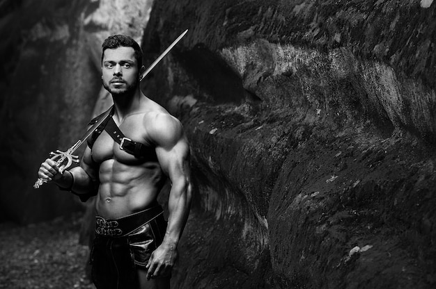 Free photo calmed warrior. monochrome shot of a handsome muscular strong young gladiator warrior holding a sword copyspace