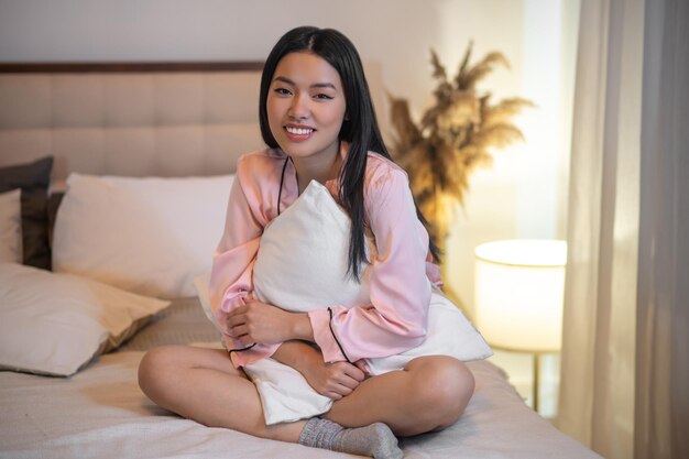 평온. 카메라를 보고 베개를 껴안고 침대에 앉아 핑크 잠옷에 긴 검은 머리를 가진 젊은 성인 웃는 아시아 여성