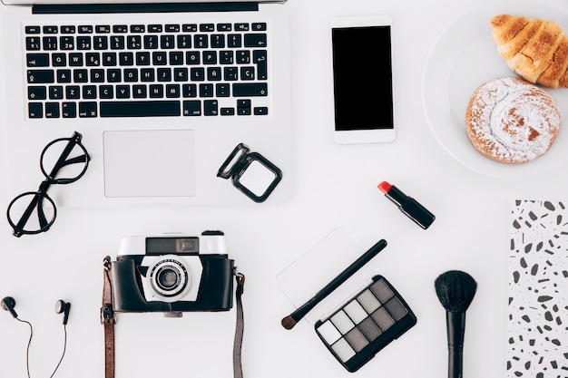 Camera; laptop; mobilephone; cosmetics product and baked pastry on white desk
