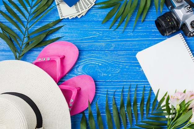 Camera near hat with flip flops and notepad among plant leaves