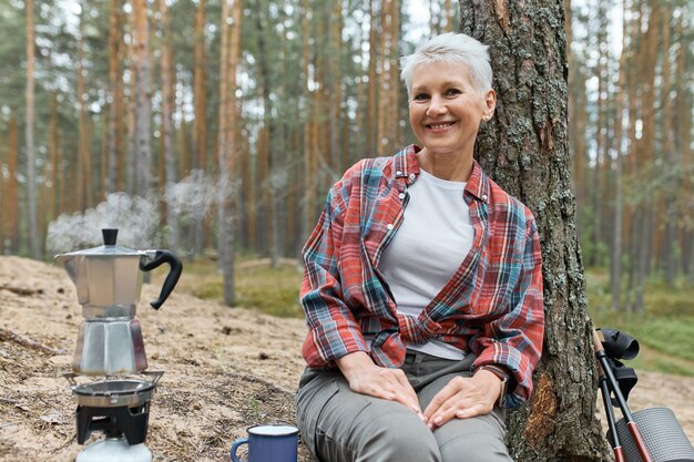 Camping lifestyle in forest. Cheerful middle aged Euroepan woman sitting on ground under pine going to make tea, boiling water in kettle on gas stove burner, having joyful happy facial expression