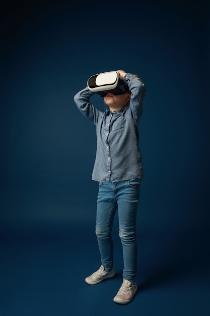 Can't believe her eyes. Little girl or child in jeans and shirt with virtual reality headset glasses isolated on blue studio background. Concept of cutting edge technology, video games, innovation.