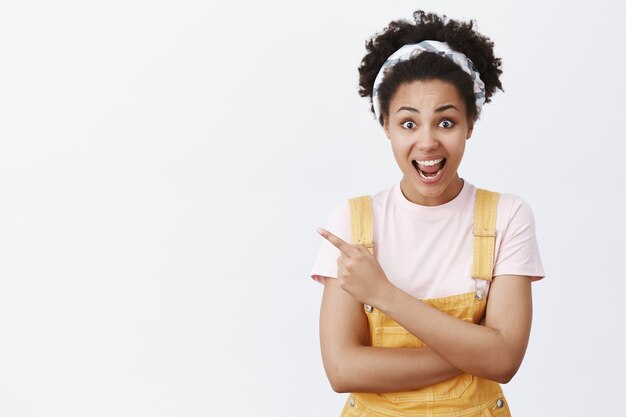 Can you believe it, I know him. Portrait of amazed excited charming dark-skinned female in headband and overalls, pointing left and smiling broadly while sharing emotions and impressions with friend